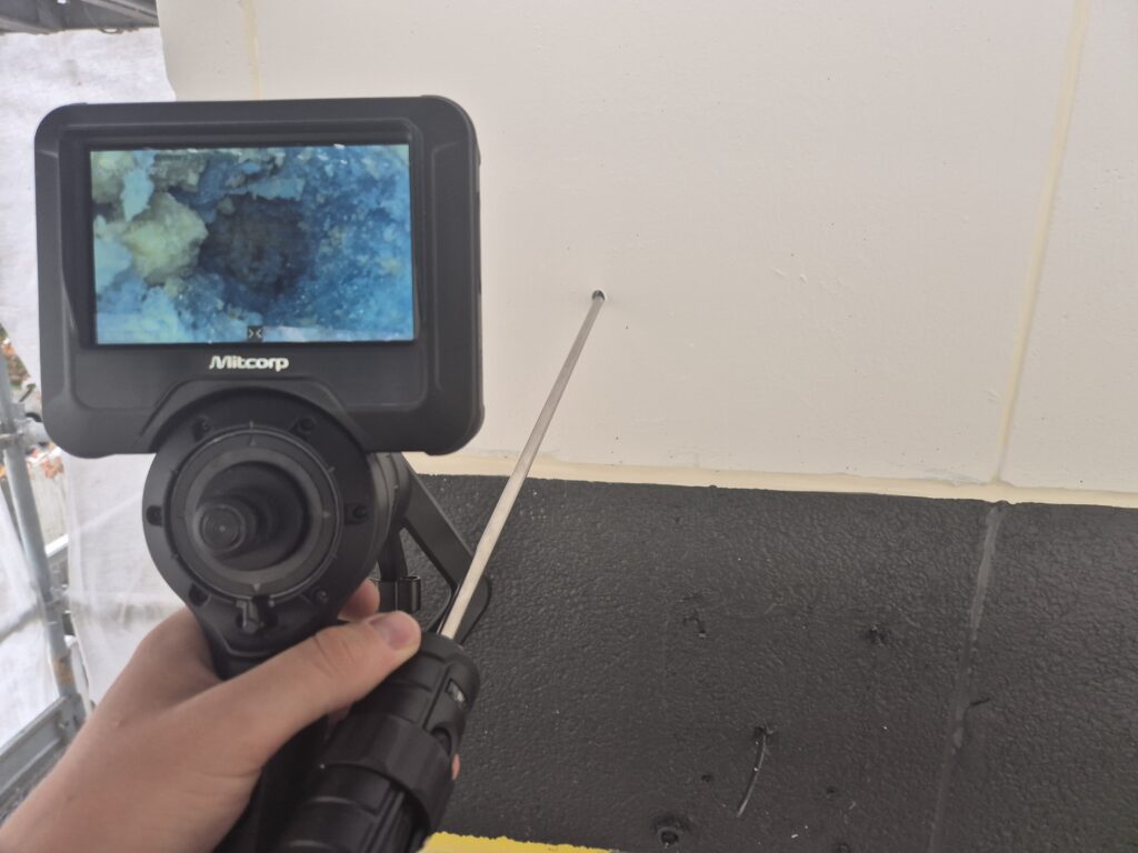 Een hand houdt een inspectiecamera vast met een scherm waarop een vergrote afbeelding van een wand te zien is. Een dunne staaf is tegen de wand aangedrukt. De wand is wit en de ondergrond is zwart.